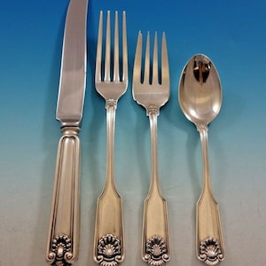 May include: A set of four silver-colored utensils: a knife, two forks, and a spoon. The handles have a decorative shell design at the base. The knife blade is smooth and the spoon has a rounded bowl. The background is a gradient of blue.