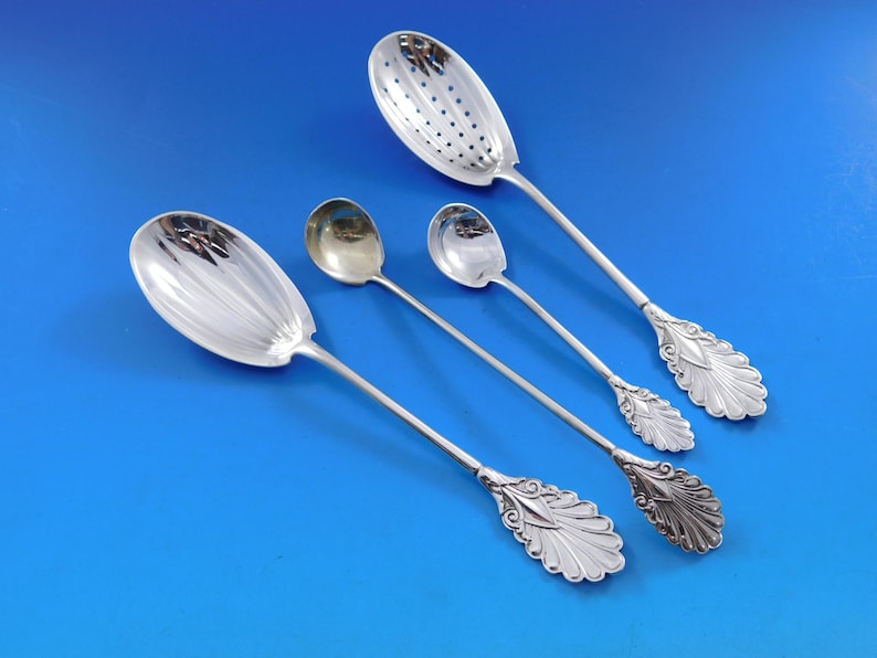 May include: Four silver spoons with ornate handles. The spoons have different shapes, including a slotted spoon and a spoon with a rounded bowl. The spoons are arranged on a blue background.