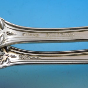 May include: Close-up of a silver serving utensil with ornate floral and leaf detailing. The word "STERLING" is stamped on the handle, along with the maker's mark and "Reed & Barton". The background is a gradient of blue.