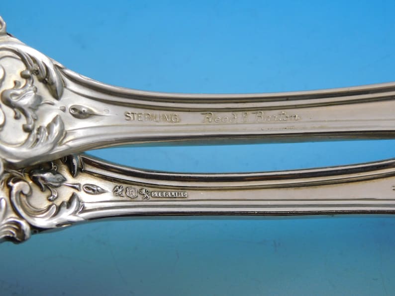 May include: Close-up of a silver serving utensil with ornate floral and leaf detailing. The word "STERLING" is stamped on the handle, along with the maker's mark and "Reed & Barton". The background is a gradient of blue.