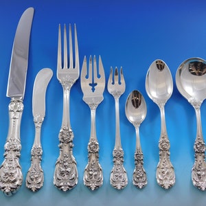 May include: An assortment of silver-coloured cutlery, comprising a knife, butter knife, forks, spoons, and a serving spoon. Each piece showcases intricate floral and scrollwork patterns on the handles, embodying a classic, elegant aesthetic. The set is displayed against a blue backdrop.