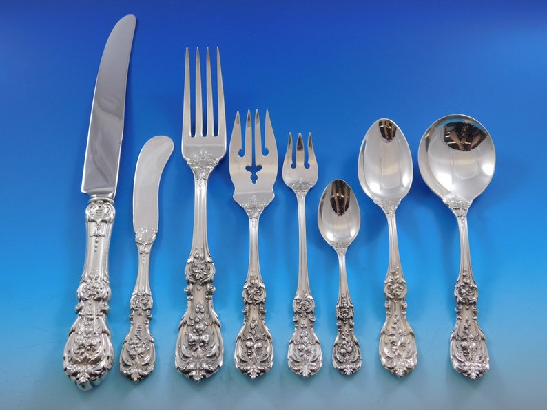 May include: An assortment of silver-coloured cutlery, comprising a knife, butter knife, forks, spoons, and a serving spoon. Each piece showcases intricate floral and scrollwork patterns on the handles, embodying a classic, elegant aesthetic. The set is displayed against a blue backdrop.