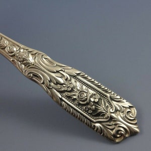 Puede incluir: Primer plano del mango adornado de una cuchara de servir de plata. El mango est&aacute; decorado con un dise&ntilde;o floral, que incluye rosas y hojas.