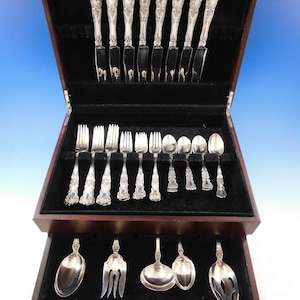 May include: A complete set of ornate silverware displayed in a dark wooden case. The set includes knives, forks, spoons, and serving utensils. The silverware has detailed floral patterns on the handles and is arranged on a black velvet lining.