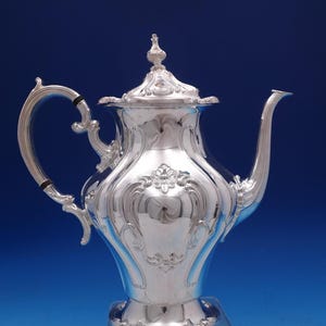 May include: A silver teapot with ornate detailing. The teapot features a curved spout, a decorative handle, and a lid with a finial. The body of the teapot has an embossed design. The background is a gradient of blue.