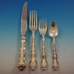 May include: A set of ornate silver flatware, including a knife, two forks, and a spoon. The handles feature a detailed, scrolling design. The knife blade is smooth and reflective. The flatware is arranged against a gradient blue and silver background.