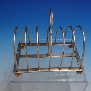 May include: A silver-toned toast rack with six arched slots and a handle. The rack has a rectangular base with rounded feet. The design is simple and elegant, suitable for serving toast at the breakfast table.