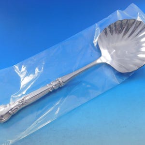 May include: A silver serving spoon with a shell-shaped bowl and an ornate handle. The spoon is packaged in a clear plastic bag.