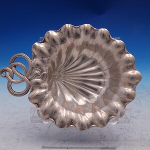 May include: A silver-colored, leaf-shaped dish with a decorative handle. The dish has a scalloped edge and a detailed, veined pattern. The handle features an intricate, looped design. The dish is likely made of metal.