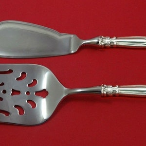 May include: A silver-colored serving set, including a pie server and a slotted serving fork, against a red background. The handles have an ornate design. The pie server has a curved blade, and the fork has a slotted design.