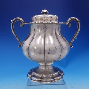 May include: A silver sugar bowl with a lid and two handles. The bowl has a rounded body and a fluted base.