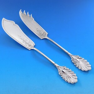 May include: Two silver butter knives with ornate handles and decorative designs on the blades. The knives have a polished finish and are in excellent condition.