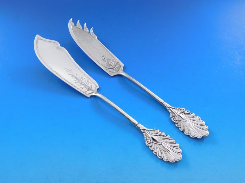 May include: Two silver butter knives with ornate handles and decorative designs on the blades. The knives have a polished finish and are in excellent condition.