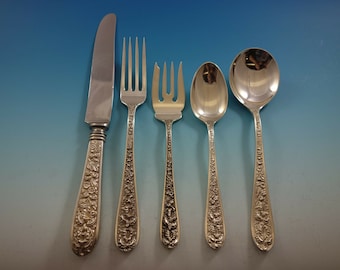 Könnte beinhalten: Ein Set aus fünf silberfarbenen antiken Besteckteilen. Das Set umfasst ein Messer, zwei Gabeln und zwei Löffel. Die Griffe sind aufwendig mit floralen Mustern verziert. Das Besteck ist vor einem blauen Farbverlaufshintergrund angeordnet.