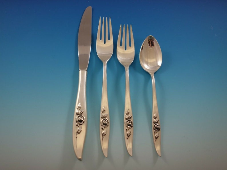 May include: A set of four silver-plated flatware pieces with rose designs on the handles. The set includes a knife, two forks, and a spoon.