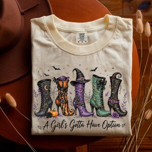 Könnte beinhalten: Ein cremefarbenes Kurzarm-T-Shirt mit einer Grafik von fünf verschiedenen Hexenstiefeln in Schwarz, Orange, Lila und Grün. Das Design enthält Fledermäuse, Sterne und einen Kürbis, mit dem Text A Girl's Gotta Have Option unter den Stiefeln gedruckt.
