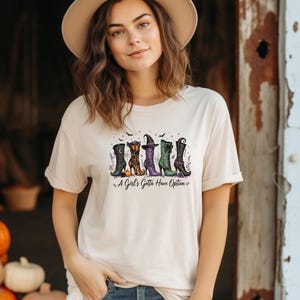 Könnte beinhalten: Ein beiges Kurzarm-T-Shirt mit einem Grafikdruck von fünf verschiedenen Hexenstiefeln in unterschiedlichen Farben und Mustern, begleitet von dem Text "A Girl's Gotta Have Option" in schwarzer Schreibschrift sowie kleinen Sternen und Fledermäusen.