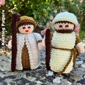 May include: Two knitted shepherd figures, one in a cream robe with a brown staff and hat, the other in a similar outfit. The figures have simple facial features and are standing on a stone surface. The words "Knitting Pattern" are in the image.