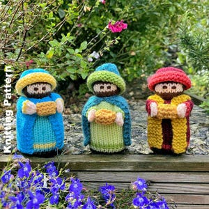 Może przedstawiać: Trzy dziergane figurki Trzech Króli, każda w innym kolorze szaty i kapelusza. Figurki trzymają mały, złoty prezent. Tło przedstawia zielone liście i fioletowe kwiaty.