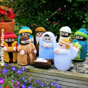 Puede incluir: Un conjunto de figuras de nacimiento tejidas a mano, que incluyen a María, José, el niño Jesús en un pesebre, un ángel y varios reyes magos. Las figuras son de varios colores, incluyendo azul, marrón, blanco y amarillo. El texto "Knitting Pattern" es visible.