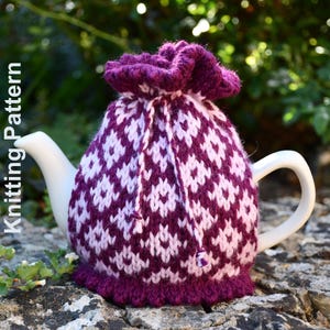 Könnte beinhalten: Eine weiße Keramikteekanne, bedeckt mit einer gestrickten Teewärmerhülle mit einem geometrischen Rautenmuster in Dunkellila und Hellrosa. Die Hülle hat ein gerafftes Oberteil mit Kordelzug und einen gerüschten unteren Rand. Text Knitting Pattern.