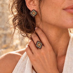 Op de afbeelding: Een vierkante oorsteker en een bijpassende ring met ingewikkeld kraalwerk. De sieraden tonen een geometrisch patroon met een centraal goudkleurig vierkant, omringd door rijen lichtblauwe en donkerbruine kralen in een ruitvormig ontwerp.