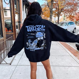 May include: A black hooded sweatshirt featuring a large graphic on the back. The design includes the text TANK GIRL in light blue, along with an illustration of a character wearing a helmet, smoking, and holding a weapon, with Alan Martin Jamie Hewlett text.