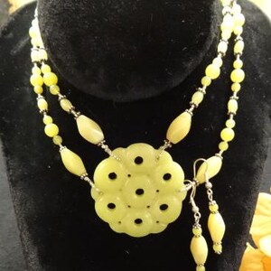May include: A double strand necklace with a large, carved jade pendant in the shape of a flower. The necklace is made of light green jade beads and silver beads. The pendant has a delicate, intricate design with small holes throughout. The necklace is paired with matching earrings.