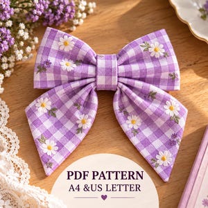 Op de afbeelding: Een stoffen haarstrik met een paars-wit vichy-ruitpatroon, geborduurd met witte madeliefjes en kleine paarse knopjes. Een rond label onder de strik toont de tekst PDF PATTERN A4 & US LETTER met een klein hartvormig icoontje.