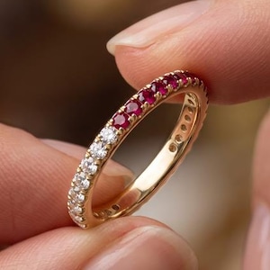 May include: A gold band ring featuring a row of alternating round-cut gemstones, with a section of clear white stones followed by a section of deep red rubies set along the top half of the metal band in a channel or prong setting style.