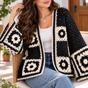 Op de afbeelding: Een zwart en crème gehaakt vest met een herhalend granny square patroon met bloemmotieven. Het kledingstuk is aan de voorzijde open, heeft lange, wijde mouwen en is opgebouwd uit garenvierkanten die afwisselen tussen zwarte en crèmekleurige centra.