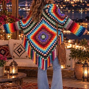 May include: A multi-colored crochet cardigan featuring a large square pattern with vibrant hues of red, orange, yellow, green, blue, and purple. The garment has long sleeves, a pointed hemline with long fringe tassels, and is worn with blue jeans.