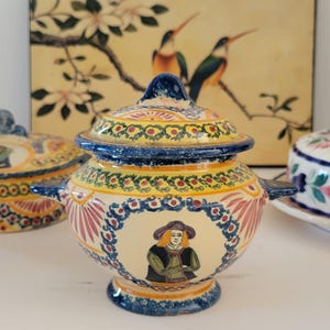 May include: A ceramic lidded sugar bowl with two handles, featuring a cream-colored base, blue and yellow floral borders, and a central hand-painted illustration of a person wearing a hat and vest, accented with red and green decorative patterns.