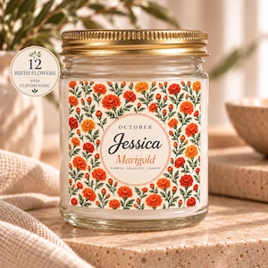 May include: A clear glass candle jar with a gold metal lid featuring a label with orange and yellow marigold flowers. The label includes the text October, Jessica, Marigold, Warmth, Creativity, Passion, and a circular badge stating 12 Birth Flowers.