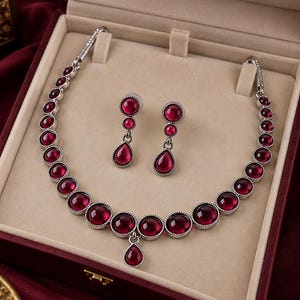 Pode incluir: Um conjunto de joias composto por um colar e um par de brincos pendentes. O colar apresenta uma série de pedras cabochão redondas de cor vermelho-escuro engastadas em metal prateado, com um pingente em forma de gota no centro.