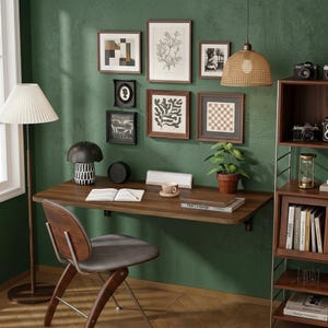 May include: A wall-mounted wooden desk with a chair, a floor lamp with a pleated shade, and a rattan pendant light. The green wall features a gallery of framed art, while a wooden shelving unit holds cameras, books, and an hourglass.