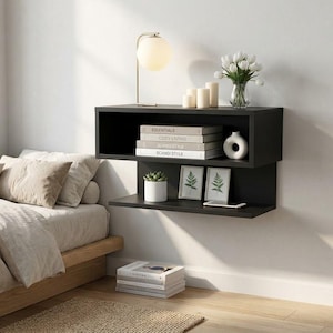 May include: A black wall-mounted floating shelf unit featuring an upper rectangular cubby and a lower open shelf. The unit holds books labeled Essentials, Cozy Living, and Scandi Style, a white vase, a potted succulent, and two framed prints.