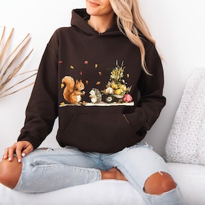 May include: A dark brown hooded sweatshirt featuring a graphic print of a squirrel and a hedgehog standing next to a small cart filled with pumpkins and corn, with scattered autumn leaves falling around them.