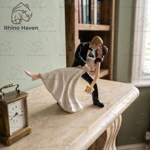May include: A wedding cake topper figurine depicting a couple in a dip pose. The figure wears a black tuxedo and the other wears a long white dress with a floral bouquet. A brass-colored analog clock sits to the left of the figurine.
