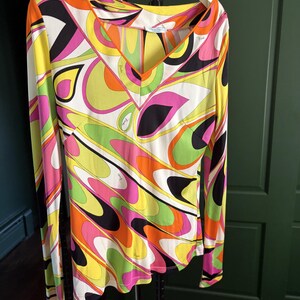 May include: A long-sleeve shirt featuring a vibrant, multi-colored abstract pattern in shades of bright yellow, lime green, orange, pink, and black. The shirt has a V-neckline with orange trim and includes the word Emilio Pucci printed on the fabric.