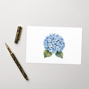 May include: A rectangular white card featuring a watercolor illustration of a blue hydrangea flower with two green leaves, positioned next to a black and gold fountain pen with a floral pattern and its matching cap placed separately nearby.