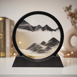 May include: A circular black frame containing a dynamic sand art display with layers of black, white, and grey sand forming mountain-like peaks and valleys. The frame is mounted on a solid black trapezoidal base, creating a stable stand.