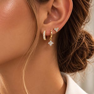 Peut inclure: Un ensemble de trois boucles d'oreilles dorées comprenant un petit anneau serti de pierres claires, un petit anneau avec une breloque fleur à quatre pétales ornée de pierres claires et une puce à trois pierres.