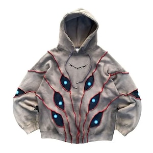 Puede incluir: Una sudadera con capucha de color topo que presenta una serie de parches oscuros en forma de ojo con centros azules brillantes. Los parches están delineados con costuras rojas prominentes que crean un efecto texturizado y superpuesto en el frente.