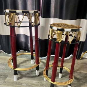 May include: Two tall stools featuring red bamboo-style legs with black and white accents. The circular seats have black and white rims, with one seat featuring a leopard print pattern. Each stool includes a gold circular footrest and support.