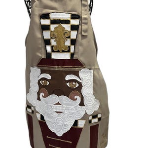 May include: A tan apron featuring a nutcracker graphic with a white pearl-embellished collar. The nutcracker has a black and white checkered hat with a gold fleur-de-lis, a white curly beard, and a dark red uniform with gold trim details.