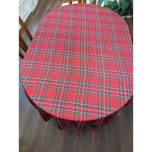 May include: An oval-shaped tablecloth featuring a classic red, green, and white plaid pattern. The fabric displays a woven texture with intersecting horizontal and vertical lines forming a traditional tartan design that drapes over the edges.