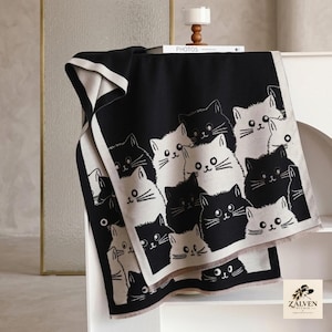 Puede incluir: Una manta de color blanco y negro con un patrón repetitivo de caras de gato estilizadas. El diseño incluye ilustraciones felinas alternas en blanco y negro con ojos grandes y bigotes, rematado con un borde sencillo a lo largo de los lados.