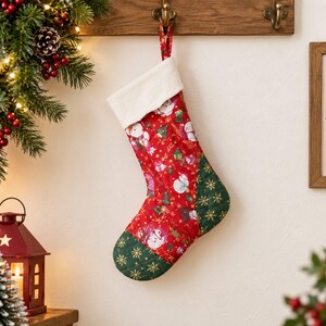 Peut inclure: Une chaussette de Noël matelassée avec un corps en tissu rouge orné d'un motif répétitif de bonhommes de neige et de sapins, un talon et une pointe vert foncé matelassés avec des flocons dorés, et un revers en peluche blanche.