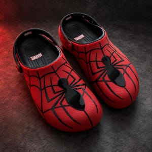 Könnte beinhalten: Ein Paar rote Clogs mit schwarzem Spinnennetz-Muster und einem markanten schwarzen Spinnen-Emblem auf der Oberseite. Das schwarze Fußbett ist mit dem weißen Marvel-Logo bedruckt. Die Fersenriemen sind schwarz und tragen ebenfalls das Marvel-Logo.